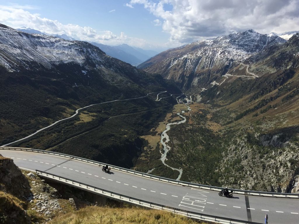 View down onto a Swiss road with views of a winding river and mountain peaks in the distance.